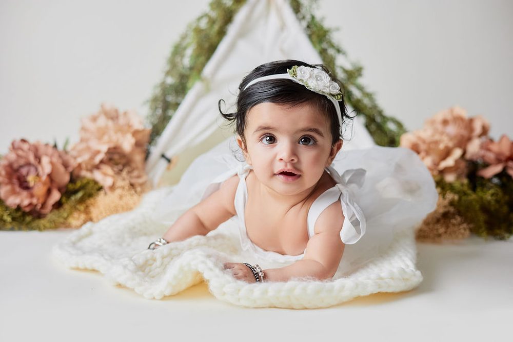 Baby girl propped on her arms on a knit blanket, dried peach florals on either side of a greenery teepee