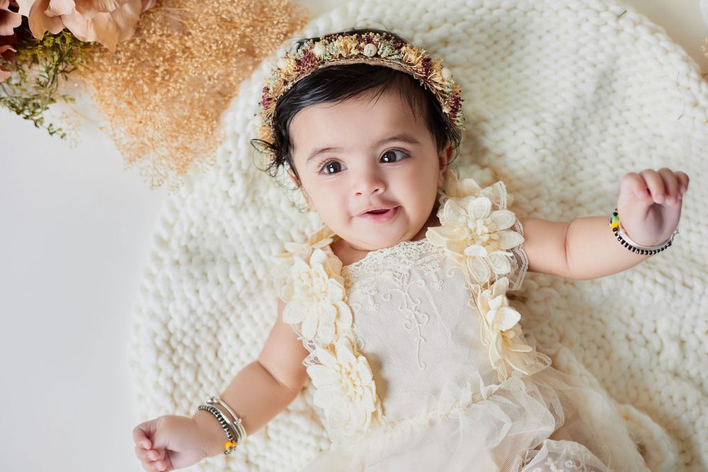 Baby in a cream lace floral dress and dried-flower crown lying on a knit blanket, smiling with an arm raised