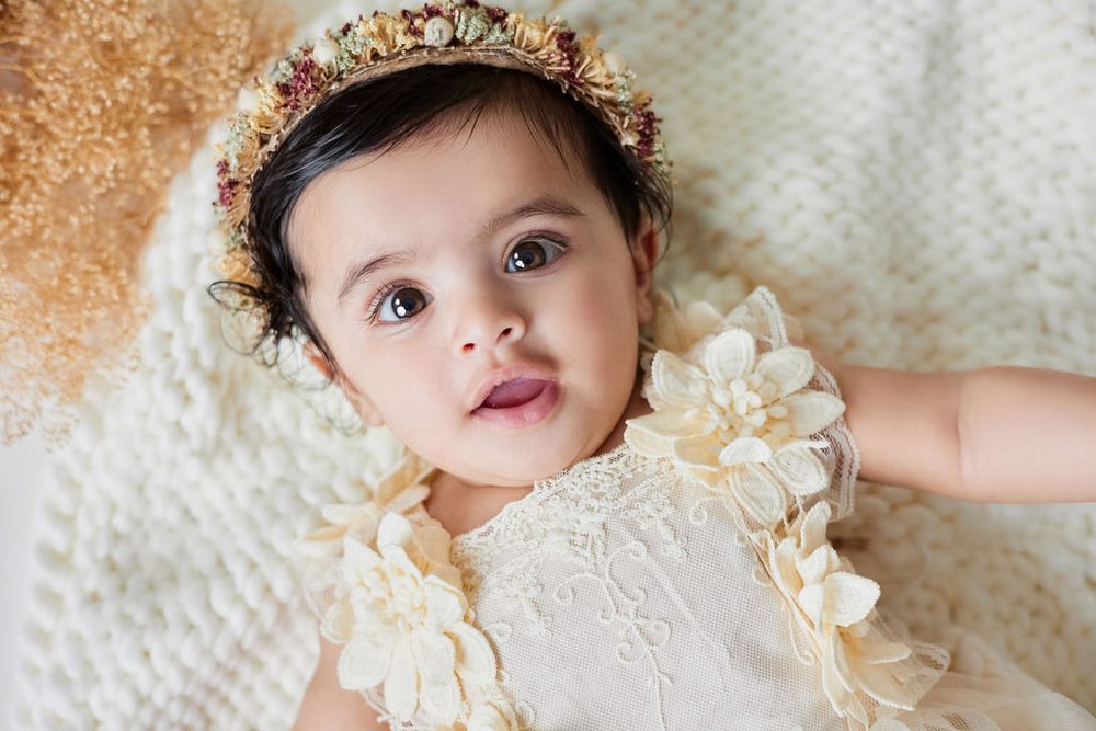 Close-up of a baby in a dried-flower crown and lace dress gazing up from a cream knit blanket