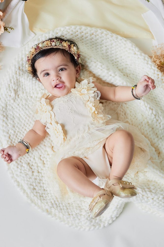 Overhead view of a baby in a lace romper and gold shoes kicking her legs on a round knit blanket