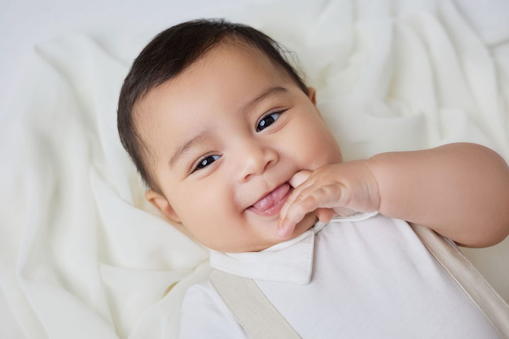 Smiling baby boy in a white collared romper chewing on his fingers while lying on soft cream fabric