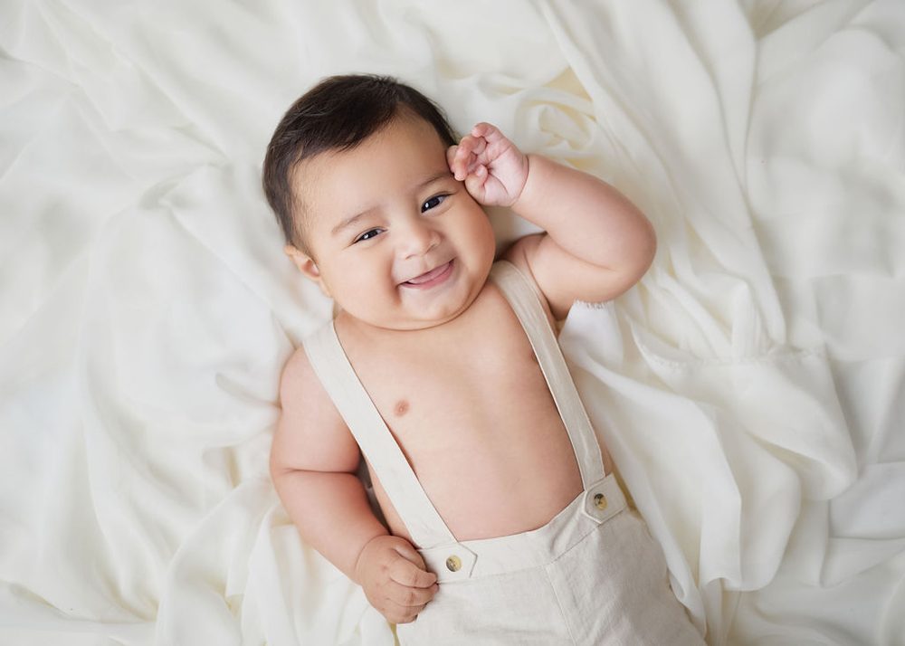Grinning baby in linen suspender pants lying on draped white fabric with a hand by his ear