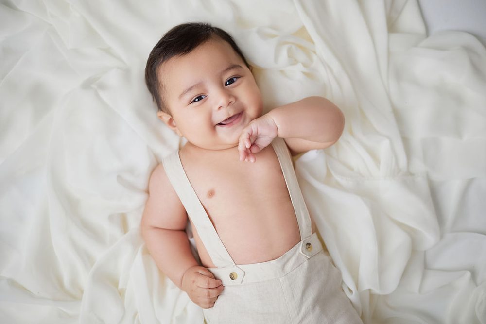 Happy baby in cream suspenders lying on white fabric with a hand tucked under his chin