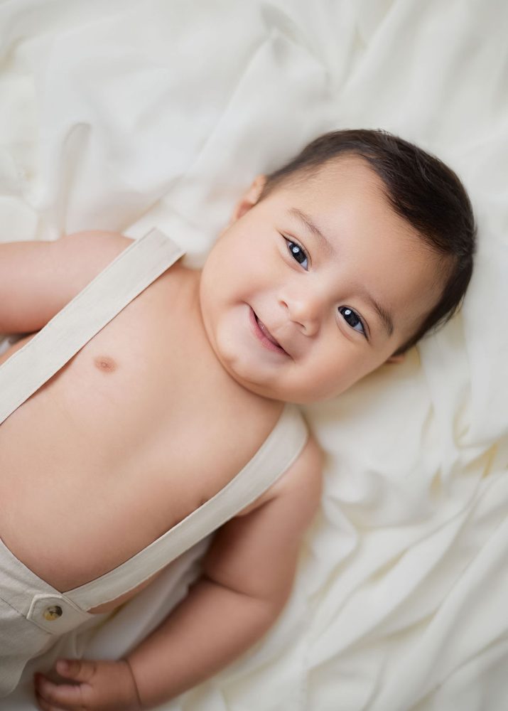 Close-up of a smiling baby boy in linen suspenders lying on rumpled ivory fabric