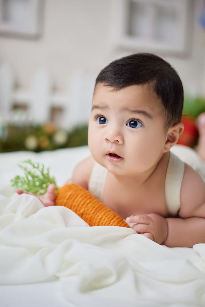 Baby on his tummy holding a knitted carrot toy on white fabric in a spring garden set
