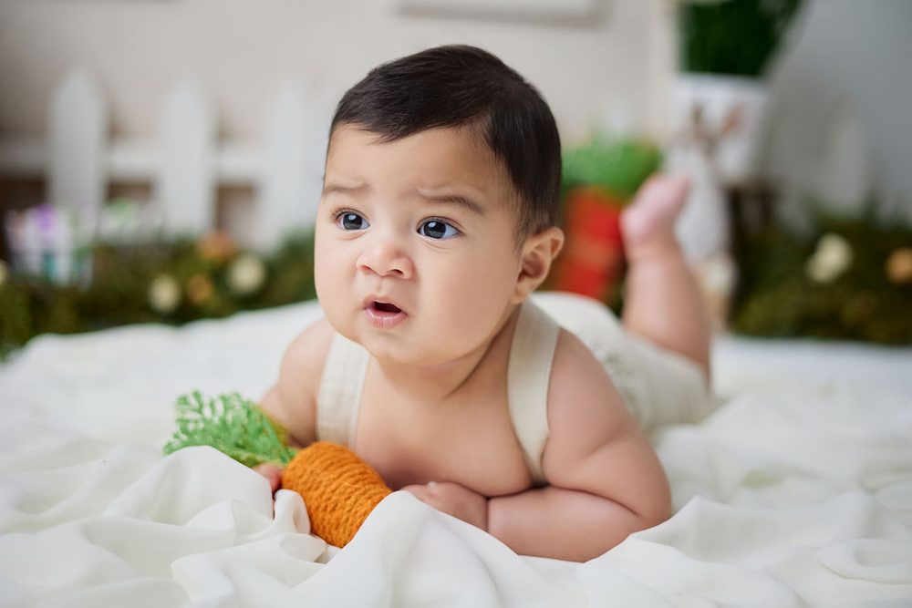 Baby propped on his forearms with a knit carrot, feet kicked up, blurred Easter set behind