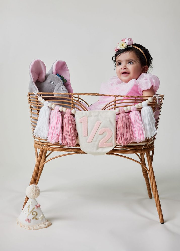 Baby girl in a pink dress sitting in a rattan bassinet with a '1/2' banner, pink tassels and plush elephant
