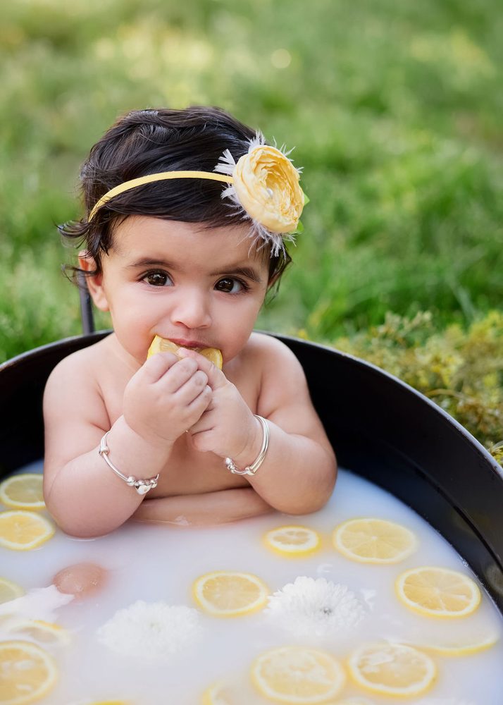 Baby in a yellow flower headband biting a lemon slice in an outdoor milk bath with floating lemons