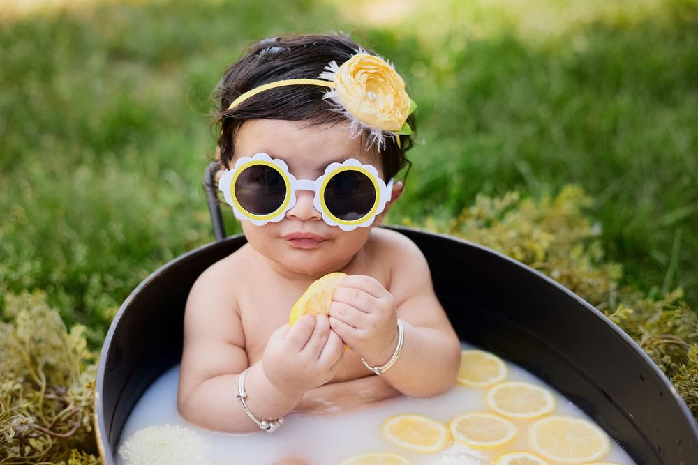 Baby in flower-shaped sunglasses holding a lemon in a milk bath tub set in the grass