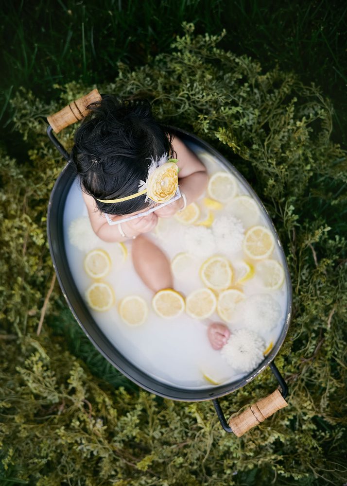 Overhead view of a baby in a metal tub milk bath with lemon slices and white mums, surrounded by greenery