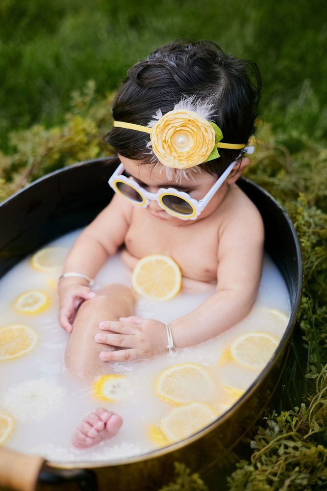 Baby in yellow-rimmed flower sunglasses looking down at lemon slices floating in her milk bath