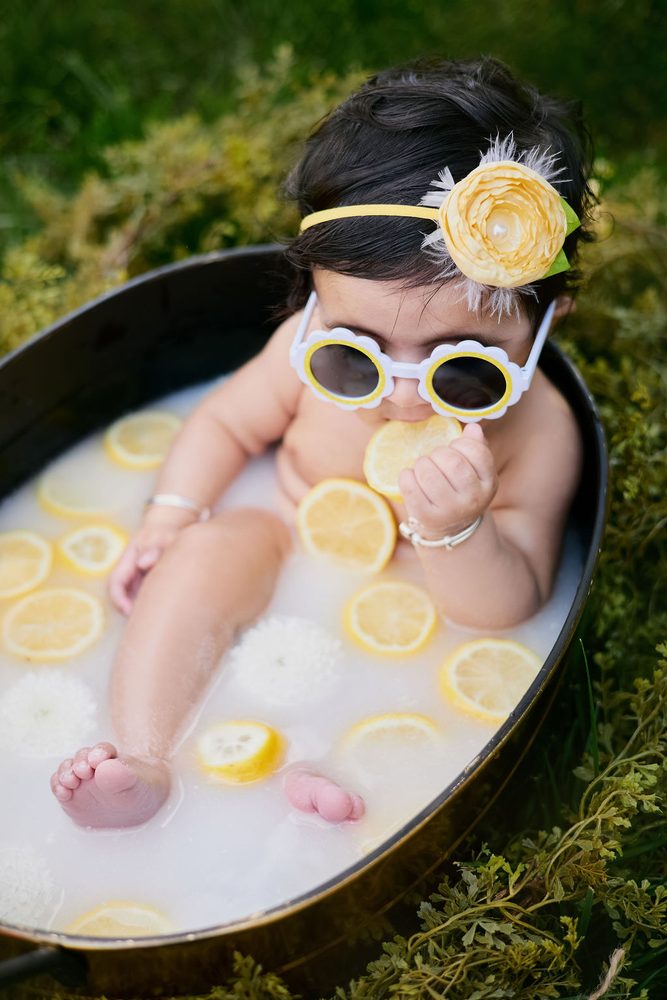 Baby nibbling a lemon slice in a milk bath, toes peeking out among floating lemons and white mums