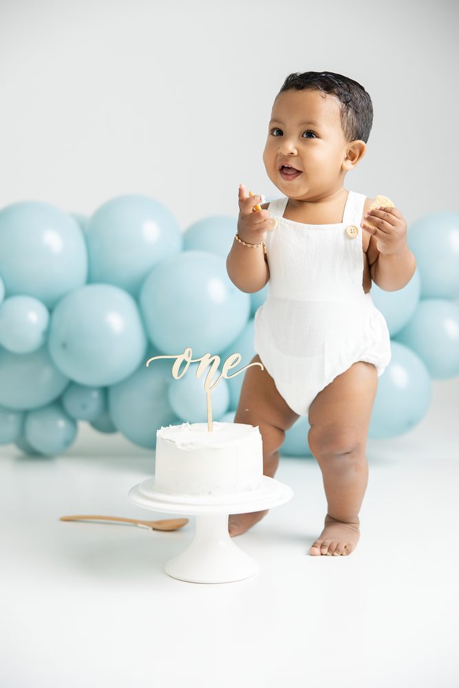Baby standing in a white romper snacking beside a white cake with a gold 'one' topper, aqua balloon garland