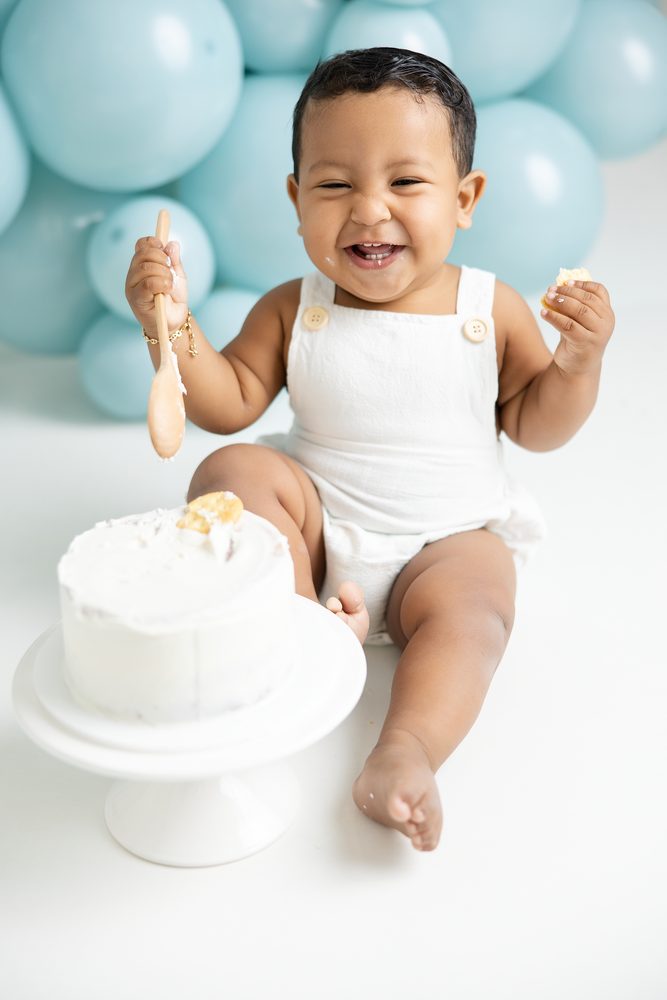 Laughing baby in a white romper waving a wooden spoon next to a white smash cake, aqua balloons behind