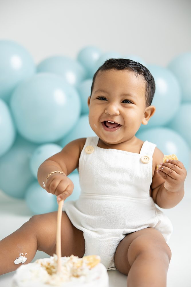 Giggling baby in a white romper with a wooden spoon in the cake and a crumb in hand, aqua balloons behind