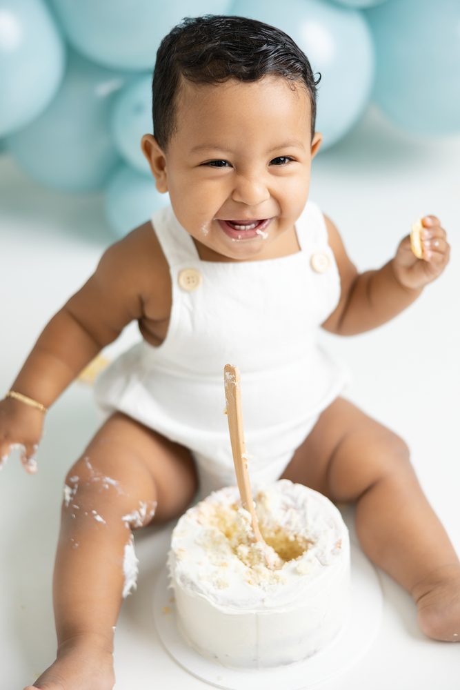 Grinning baby with frosting on his legs, a wooden spoon standing in a dug-out white smash cake