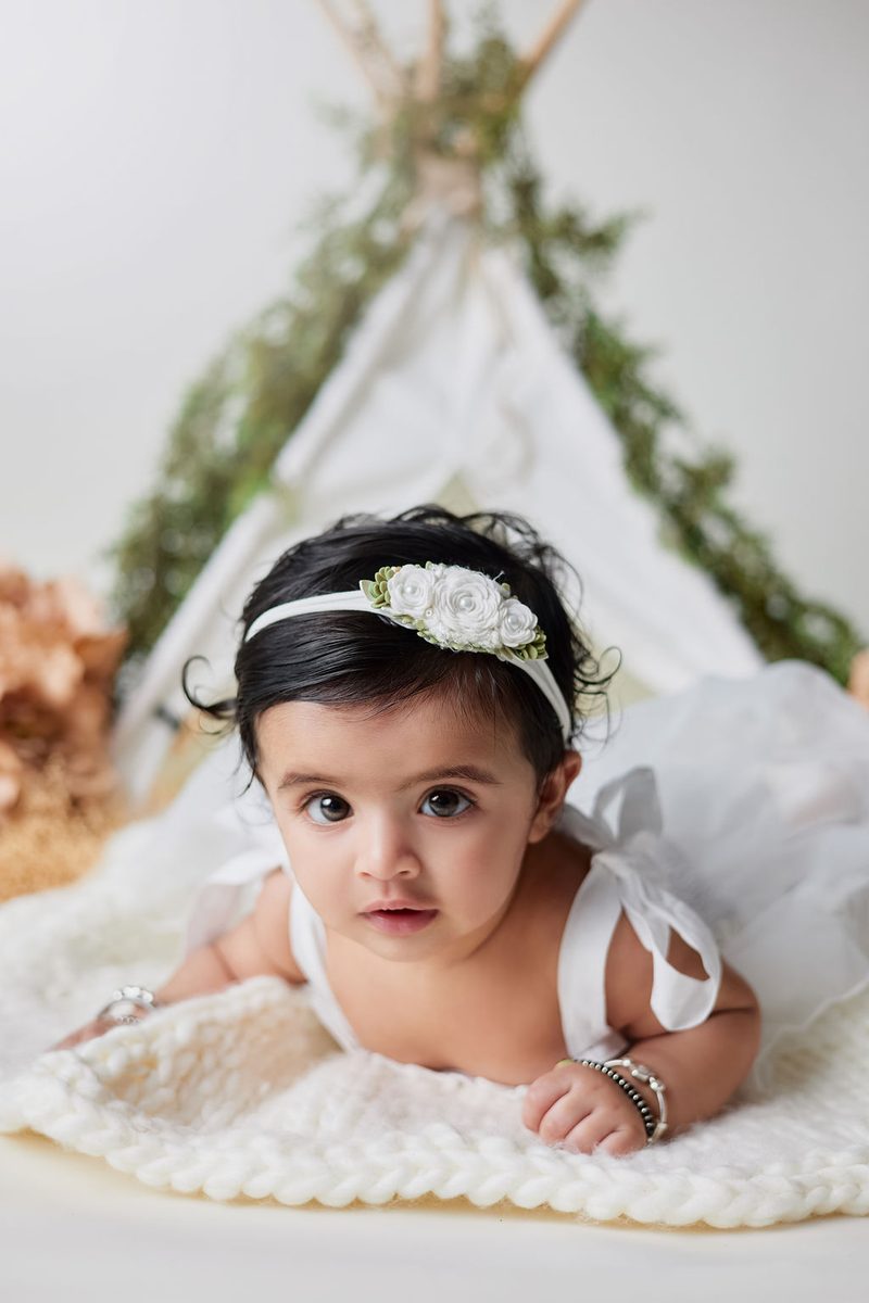 Baby milestone photography: baby girl in a rose headband on her tummy on a knit blanket before a white teepee