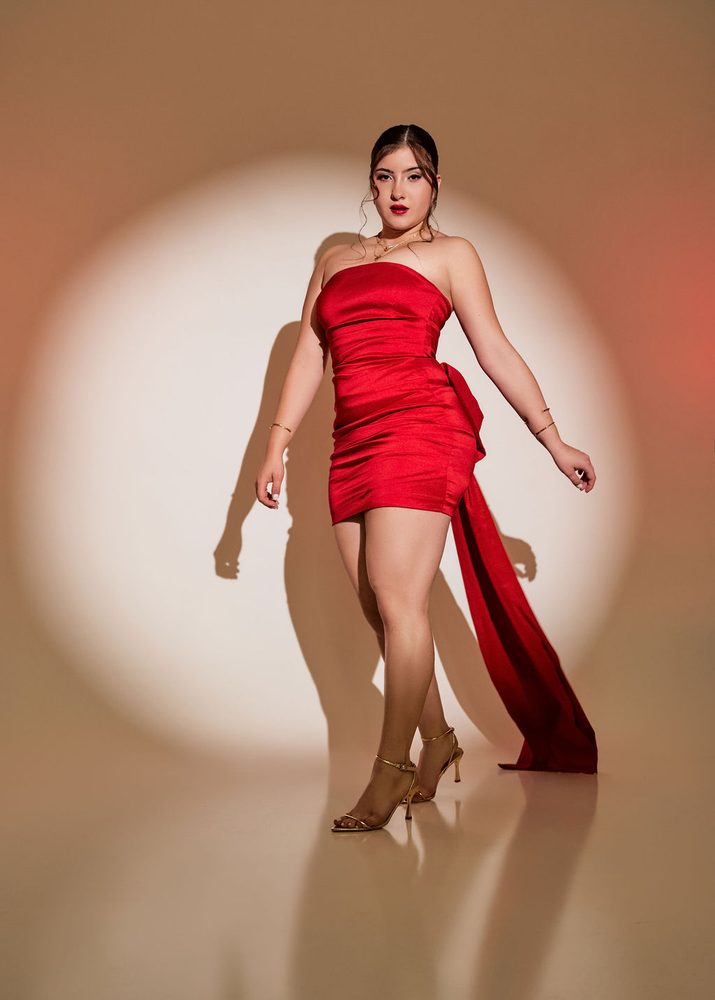 Young woman in a strapless red satin mini dress with a long bow train, posed in a circular spotlight