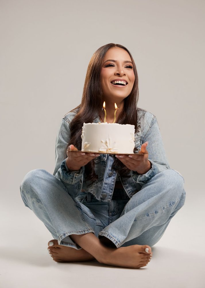 Laughing woman in a denim jacket and jeans holding a white birthday cake with two gold candles on a gray backdrop