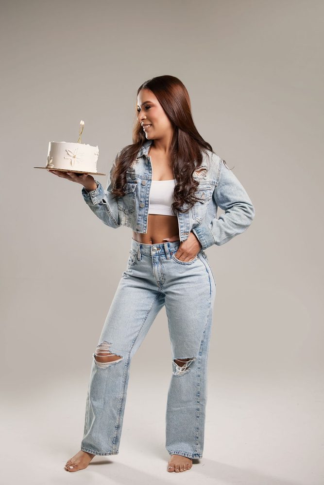 Barefoot woman in ripped jeans and a cropped denim jacket balancing a birthday cake on one hand, smiling at it