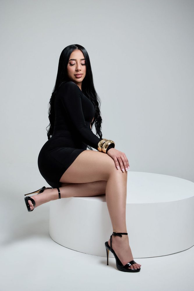 Woman in a black long-sleeve mini dress and gold cuff bracelets perched on a round white studio pedestal
