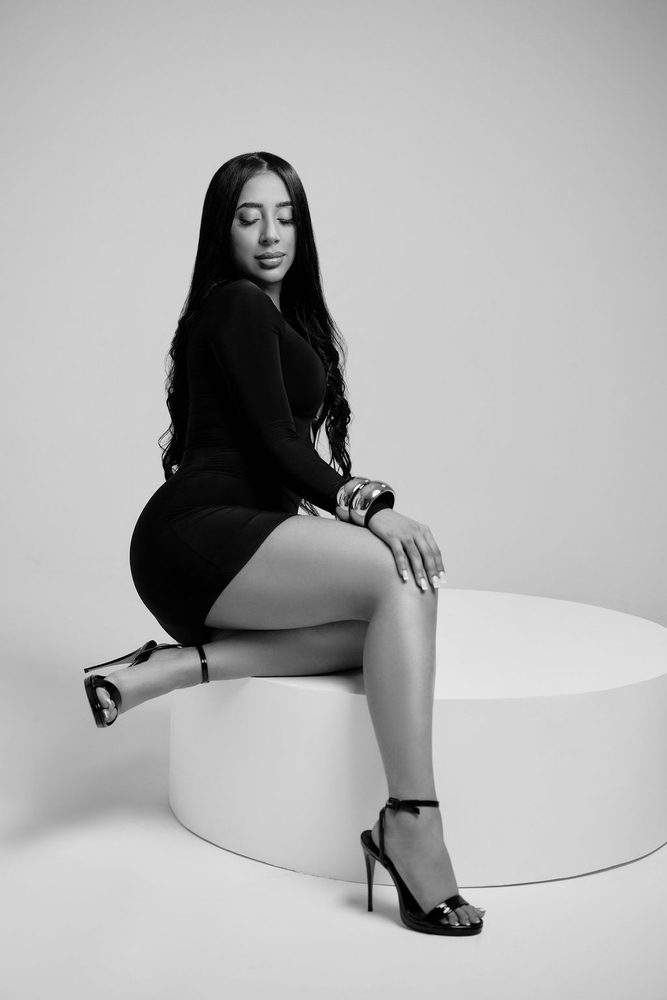 Black-and-white portrait of a woman in a fitted black dress and strappy heels seated on a round white pedestal
