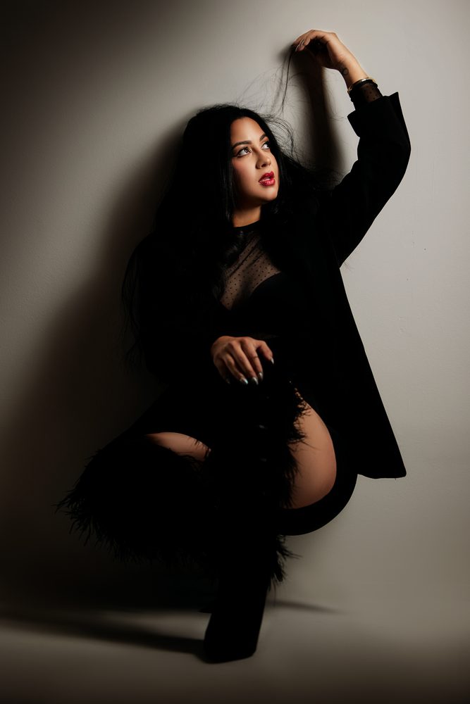 Woman crouching against a wall in a black blazer and feather boots, lifting a strand of hair in dramatic light