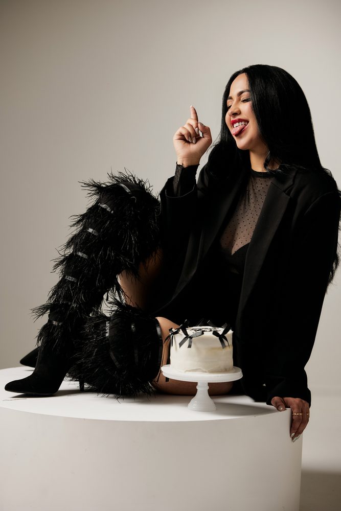 Playful woman sticking out her tongue with a finger raised, seated beside a small white cake with black bows