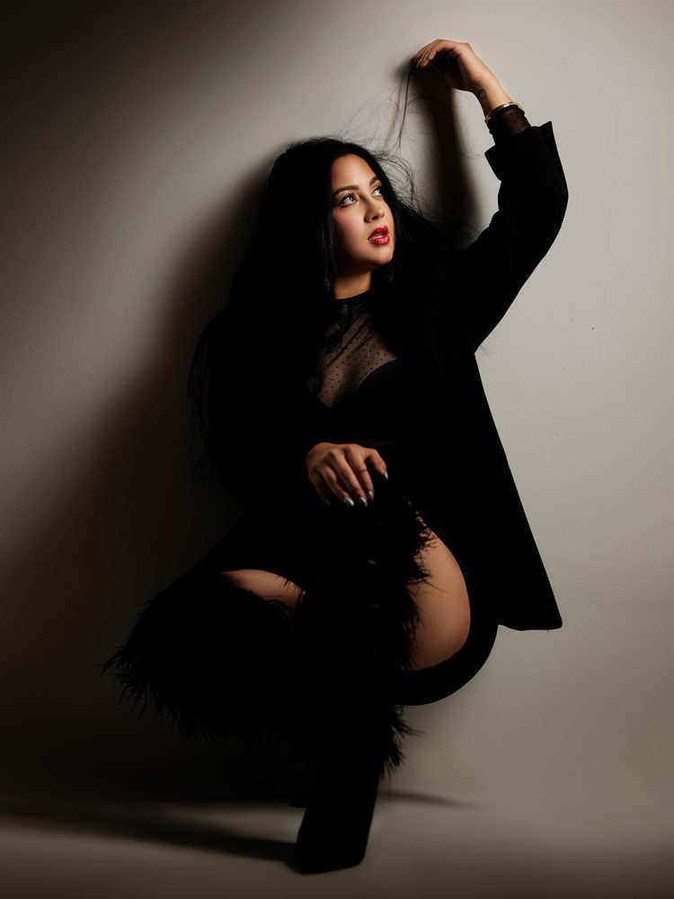 Woman with red lips crouched against a gray wall, one arm raised into her long dark hair, in black feather boots
