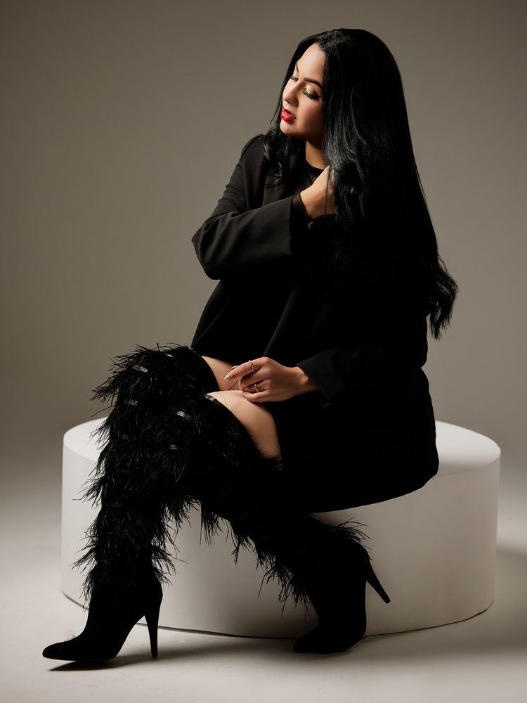 Woman in a black blazer and feathered thigh-high heels seated on a white cylinder, looking down over her shoulder