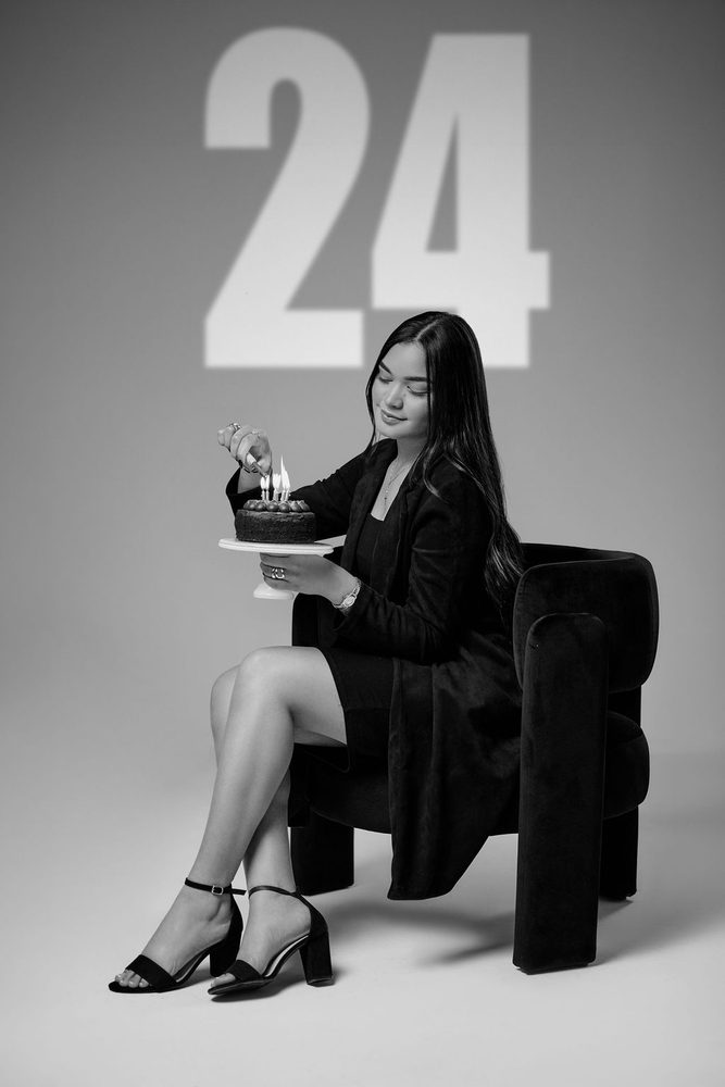 Black-and-white shot of a woman lighting birthday candles beneath a projected '24' on the studio wall