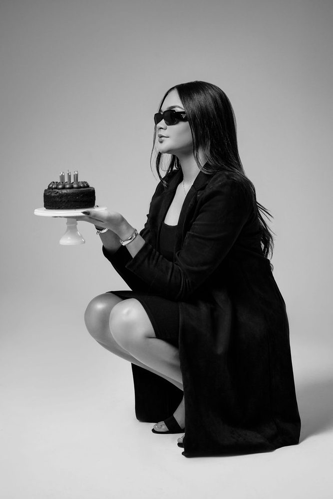 Monochrome portrait of a crouching woman in sunglasses presenting a chocolate birthday cake on a stand