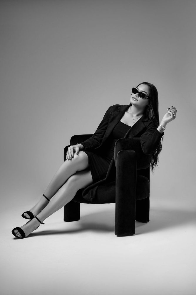 Black-and-white image of a woman in dark sunglasses reclining in a sculptural armchair, one hand raised