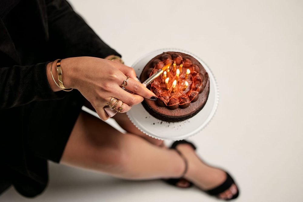 Overhead detail of a hand with gold rings lighting glowing candles on a chocolate cake resting on a white stand
