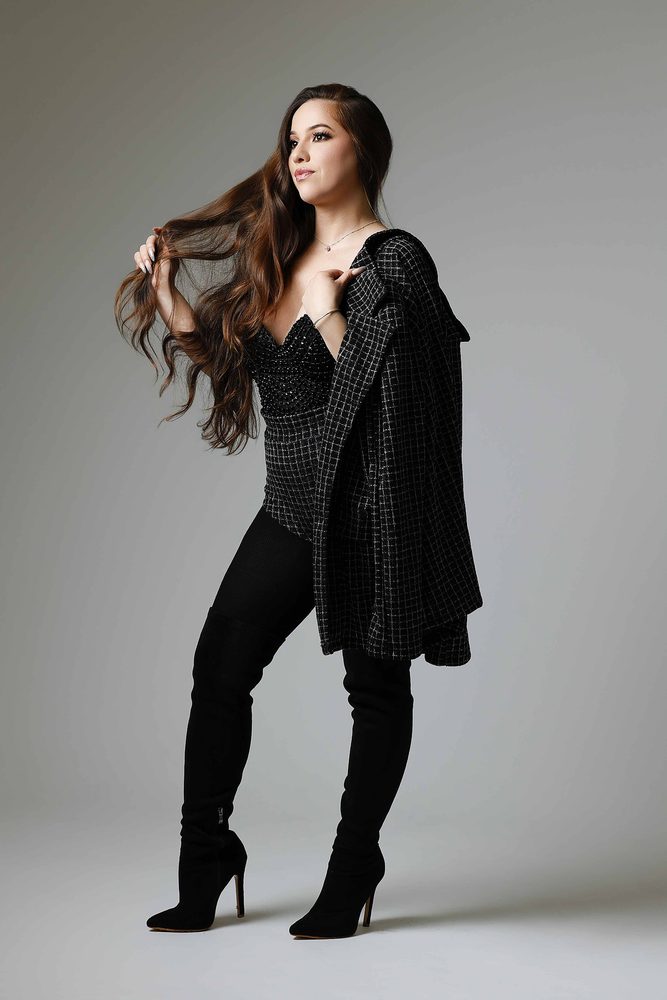 Woman in a checked black jacket draped over one shoulder and over-the-knee boots, running fingers through long hair