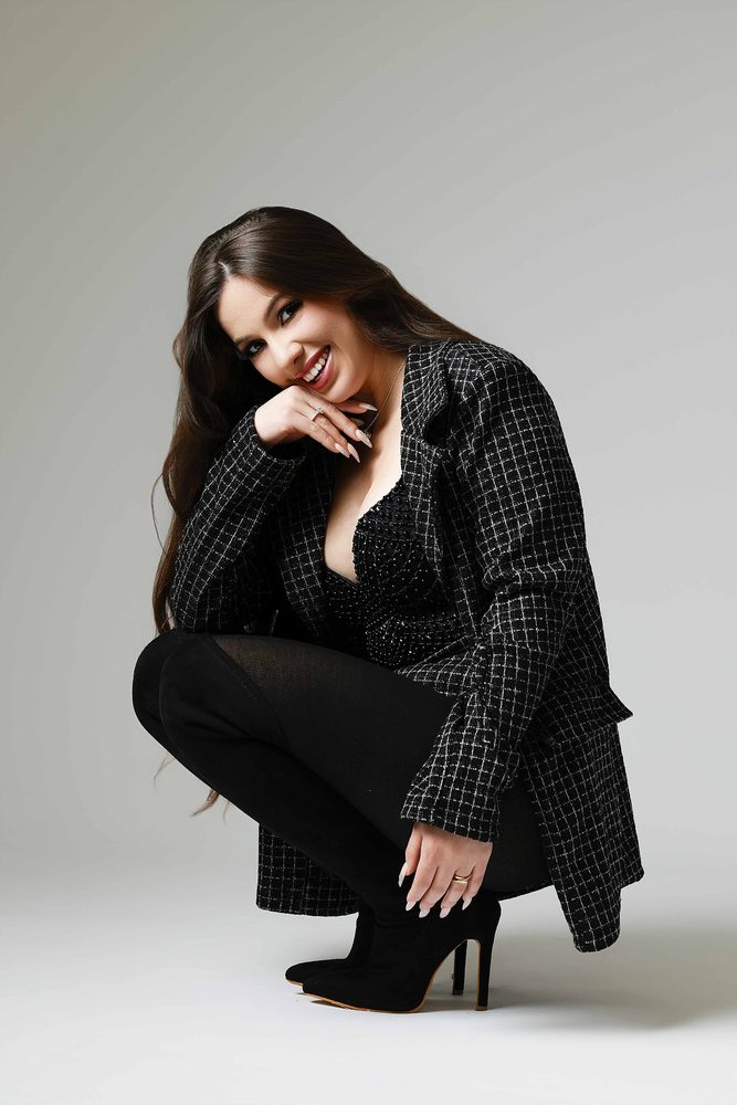 Smiling woman crouching in a black-and-white tweed coat and suede heeled boots, chin resting on her hand