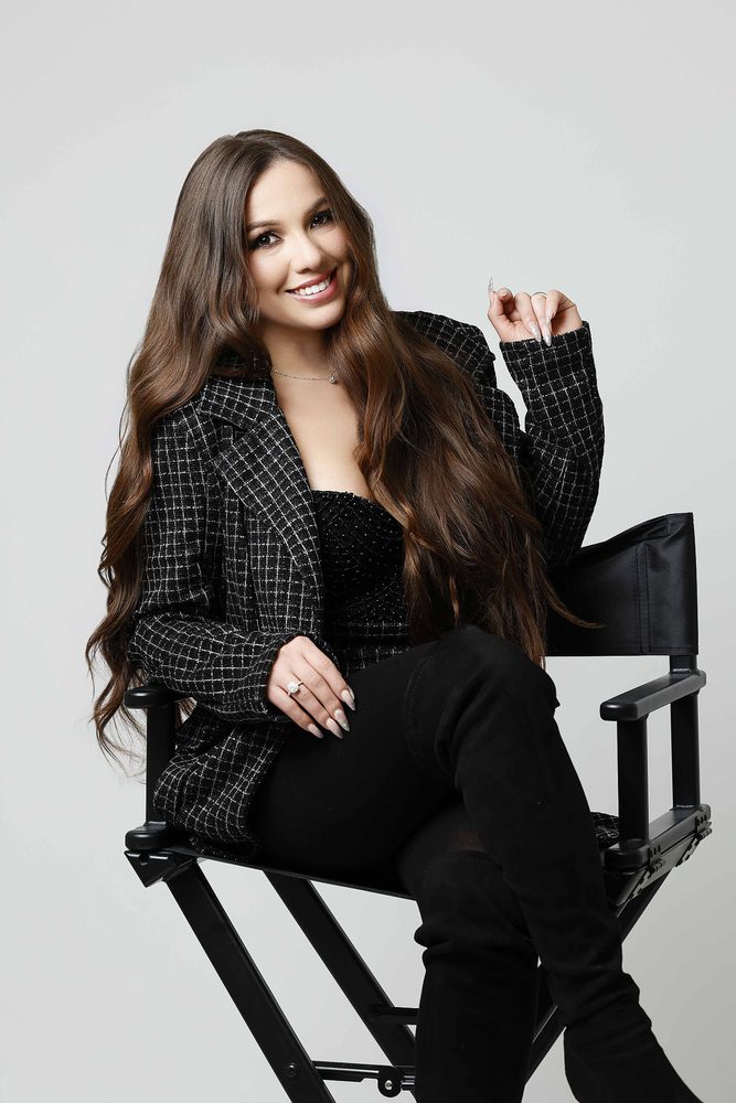 Closer shot of a smiling woman with long wavy hair in a tweed blazer, seated in a black director's chair