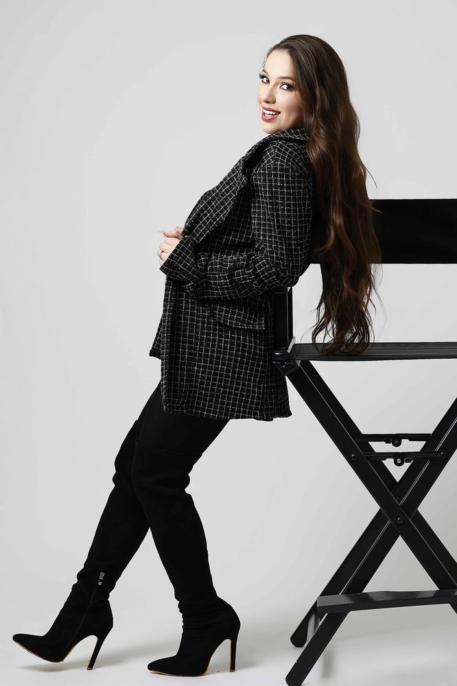Woman in a tweed coat and suede over-the-knee boots leaning back against a director's chair, smiling over her shoulder