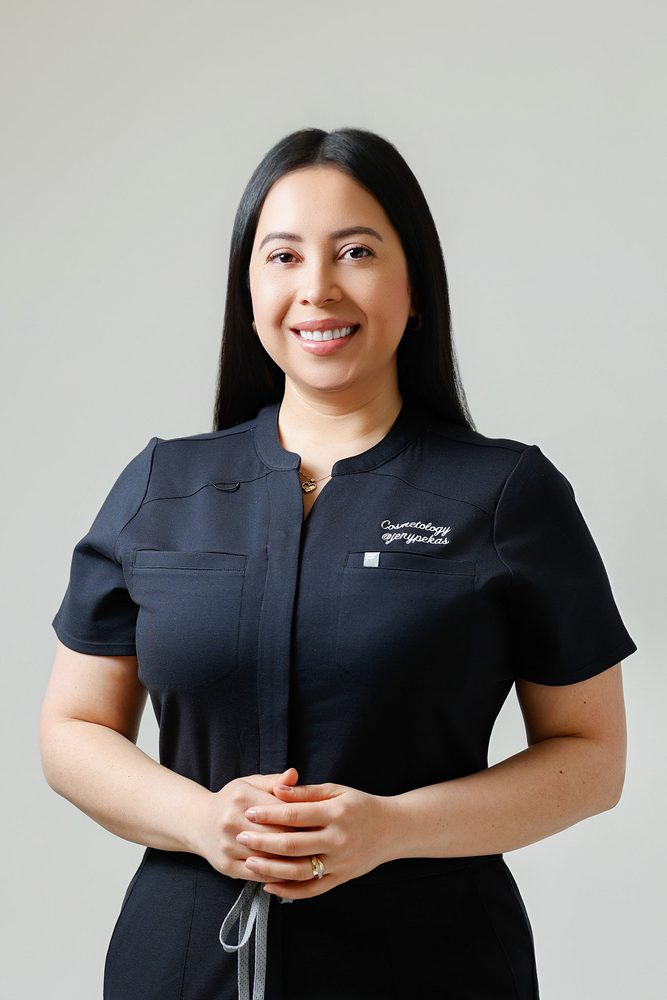 Waist-up portrait of a smiling cosmetologist with long dark hair in a black uniform, hands folded in front