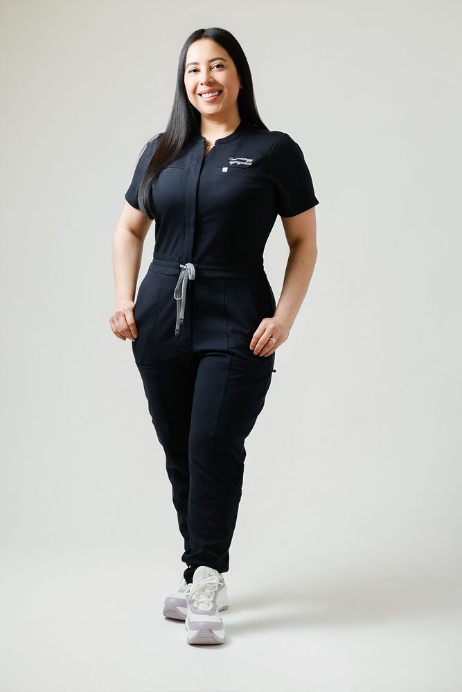 Full-length studio portrait of a woman in a black scrub jumpsuit and white sneakers, hands resting at her hips