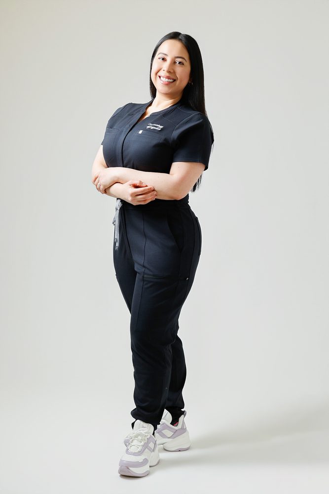 Full-length portrait of a smiling woman in black scrubs, body angled and arms crossed on a seamless gray backdrop