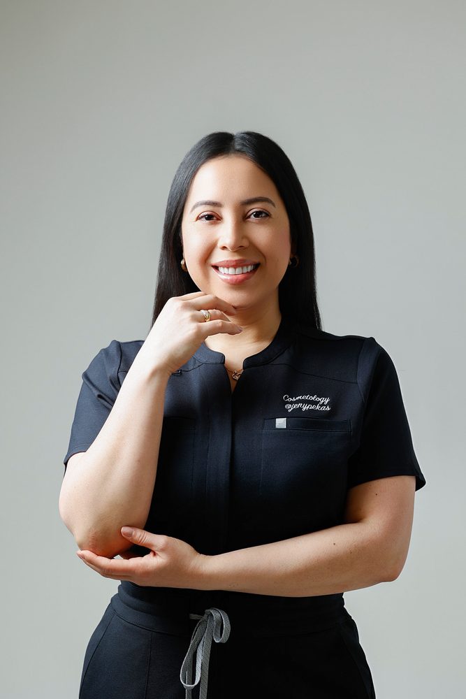 Smiling woman in black scrubs resting her chin on her hand, other arm across her waist, gray studio backdrop