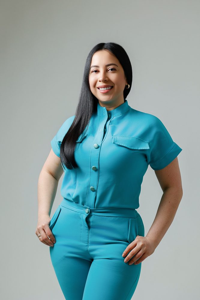 Waist-up portrait of a woman in a teal uniform with hands resting on her hips, smiling against a gray wall