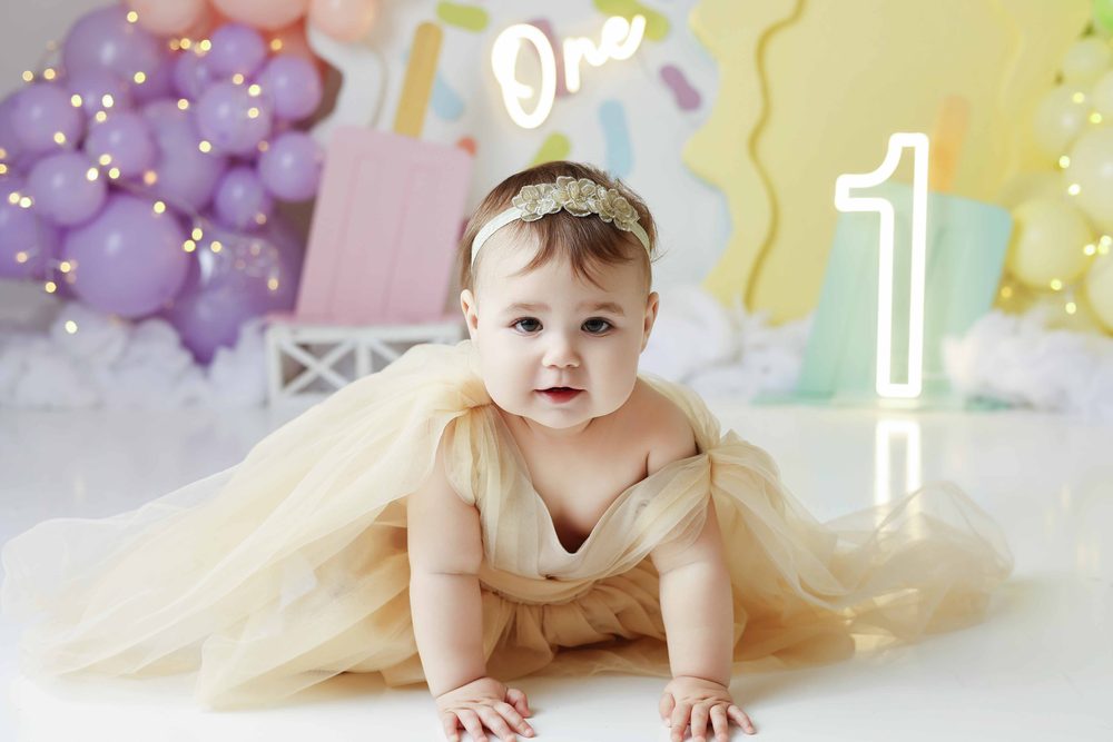 Baby girl in a cream tulle dress crawling toward the camera on a pastel popsicle set with a neon '1'