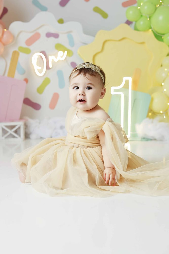 Baby girl sitting in a flowing cream tulle dress before a glowing neon 'one' sign and sprinkle backdrop