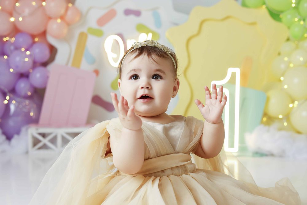 Baby clapping her hands in a cream tulle dress, purple and yellow balloon garlands with fairy lights behind