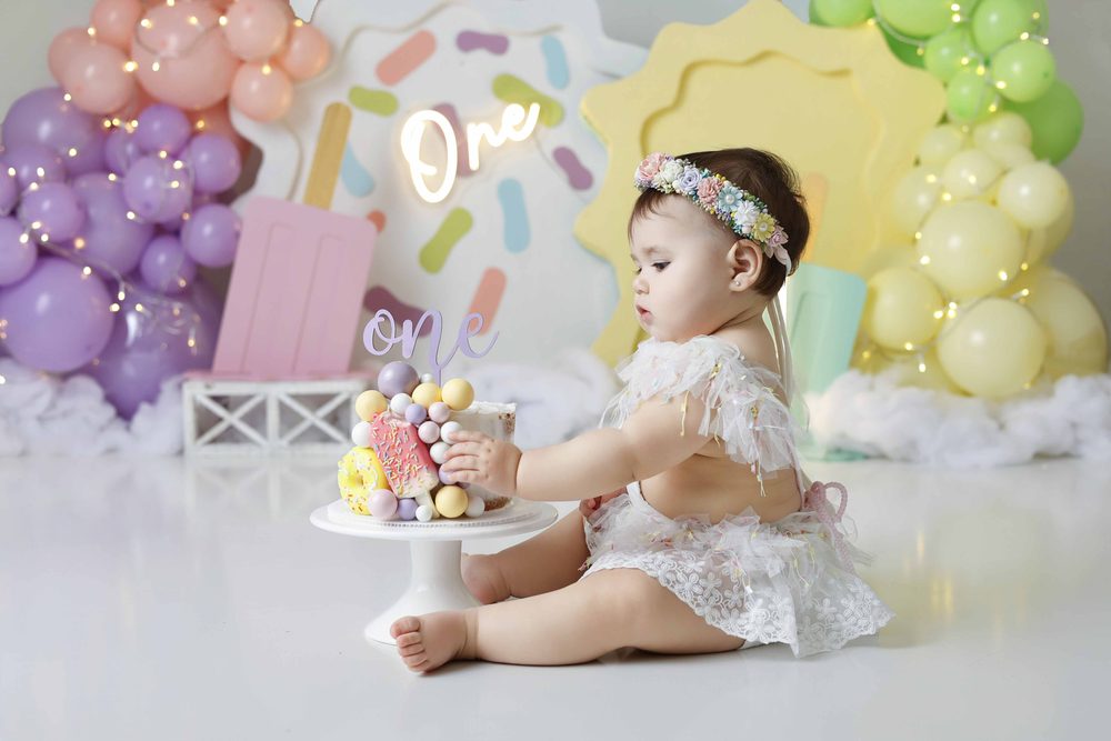 Baby in a floral crown and lace romper reaching for a sprinkle cake with candy balls and a 'one' topper