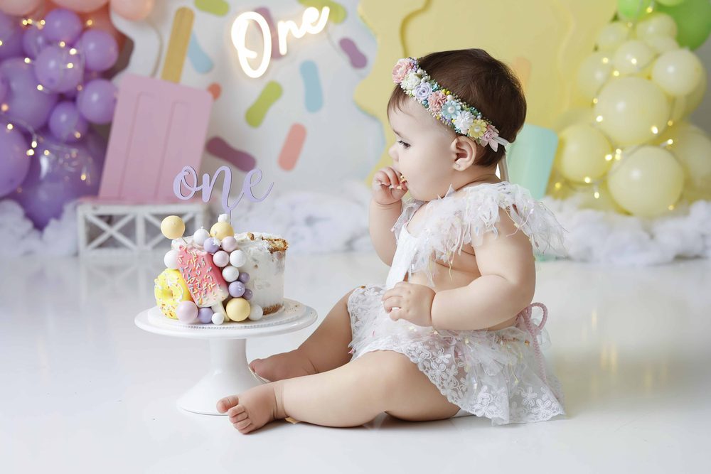 Baby tasting frosting from her fingers beside a candy-topped cake on a white stand with a 'one' topper