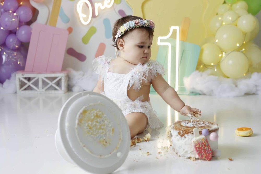 Baby grabbing chunks from a toppled smash cake, the empty cake stand tipped over in the foreground