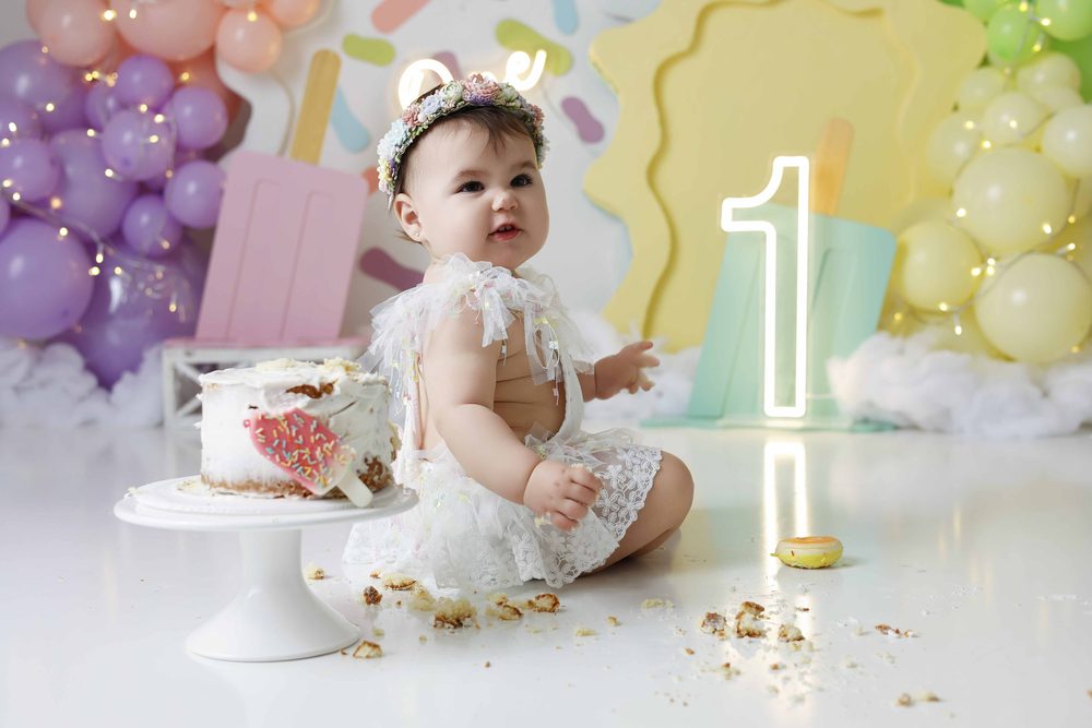 Baby in a flower crown sitting among cake crumbs beside a popsicle-topped smash cake and glowing neon '1'